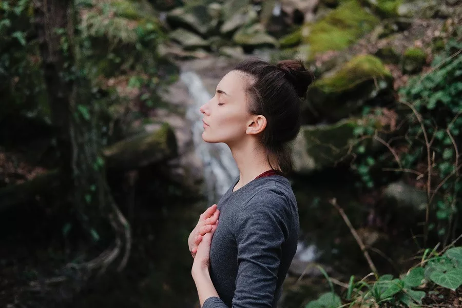 Young woman eyes closed, hands on chest, breathing outdoors in moss forest.