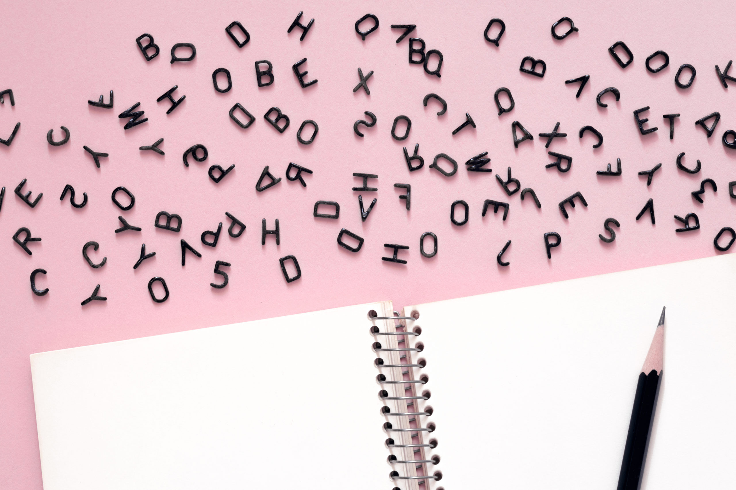 Letters of the alphabet scattered on pink surface, a clean notebook and a pencil below them.