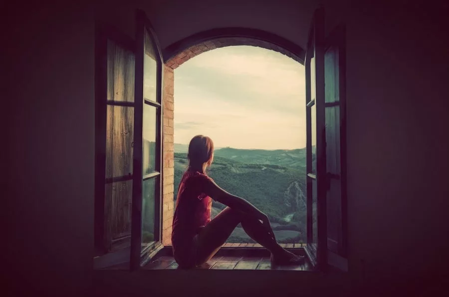 Lonely woman sitting in an open old window looking on the landscape of Tuscany, Italy.