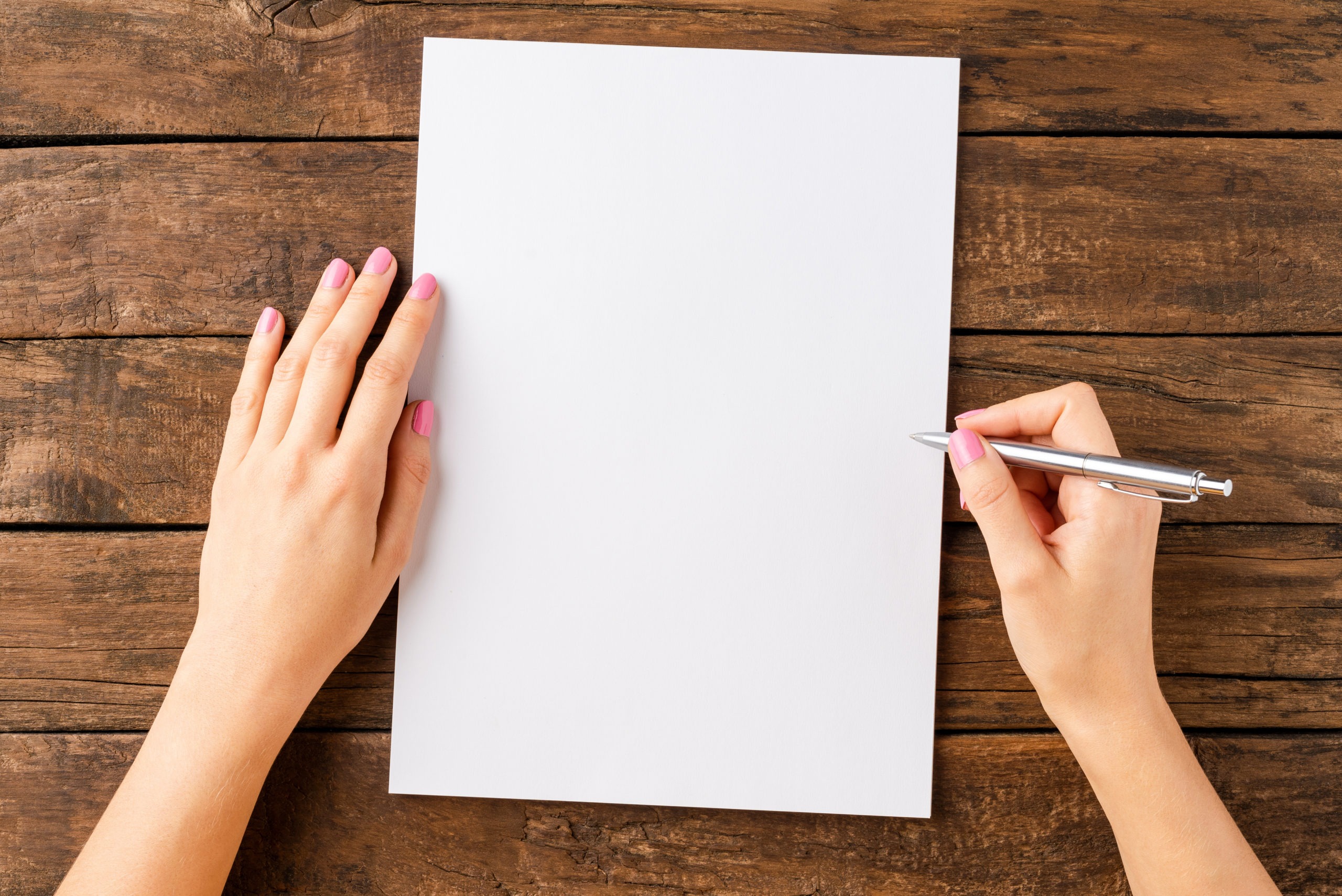 Woman’s hands writing with pen over blank white paper sheet on wooden table.