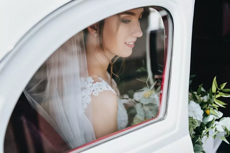 Beautiful bride with curly hair and a bouquet sits near the window in a white car.