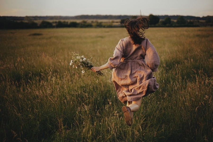 Beautiful woman in rustic dress running with wildflowers in hand in summer meadow in sunset.