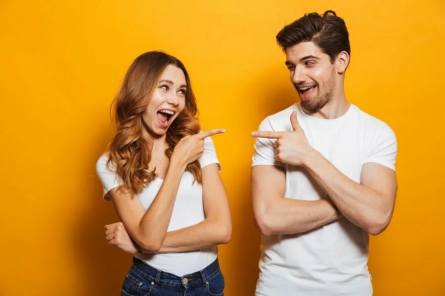 Happy young couple in basic clothing laughing and pointing fingers at each other, isolated over yellow background.