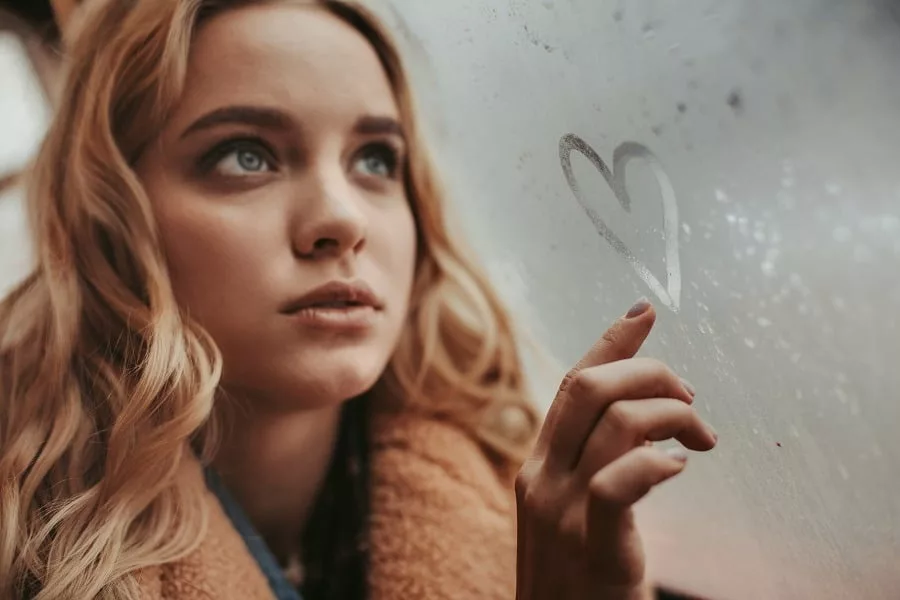Young lady with dreamy eyes drawing heart on the window.