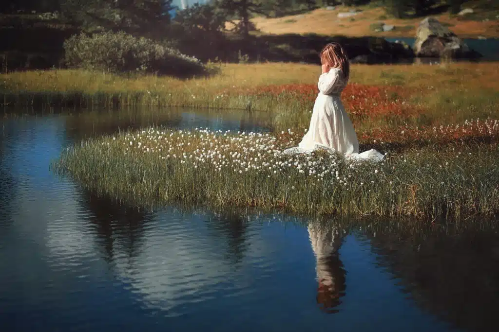 Woman with vintage dress in alpine lake kneeling and praying fervently.