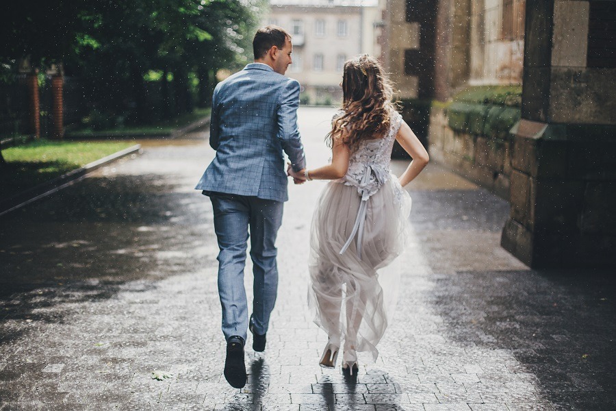 Stylish happy bride and groom running holding hands on background of old church in rainy street.