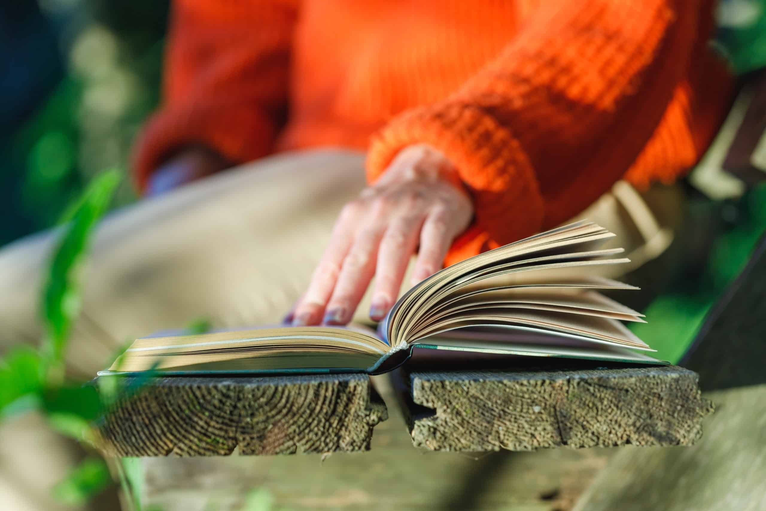 A woman leafs through the pages of a book on a wooden bench outdoor with her hand.