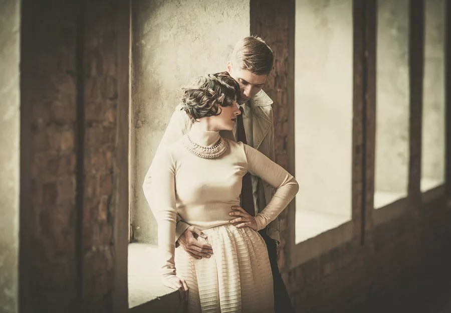 Beautiful vintage couple standing near window, woman leaning on to her man.