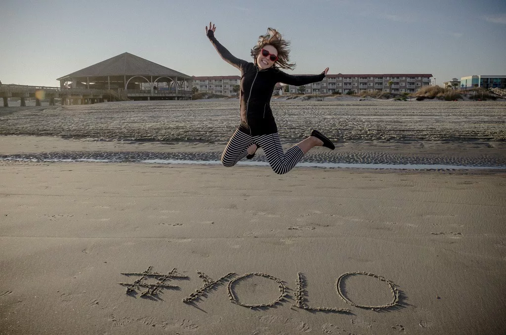 #YOLO hashtag written in the sand on the beach and an adult female jumping.
