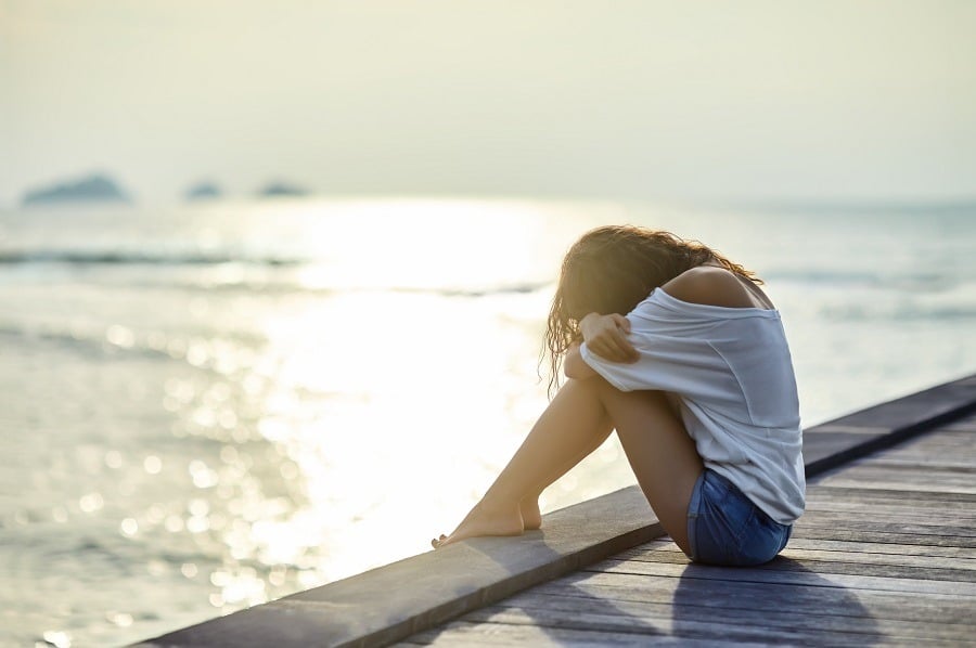 Sad lonely woman sitting on wooden dock, head tucked crying.