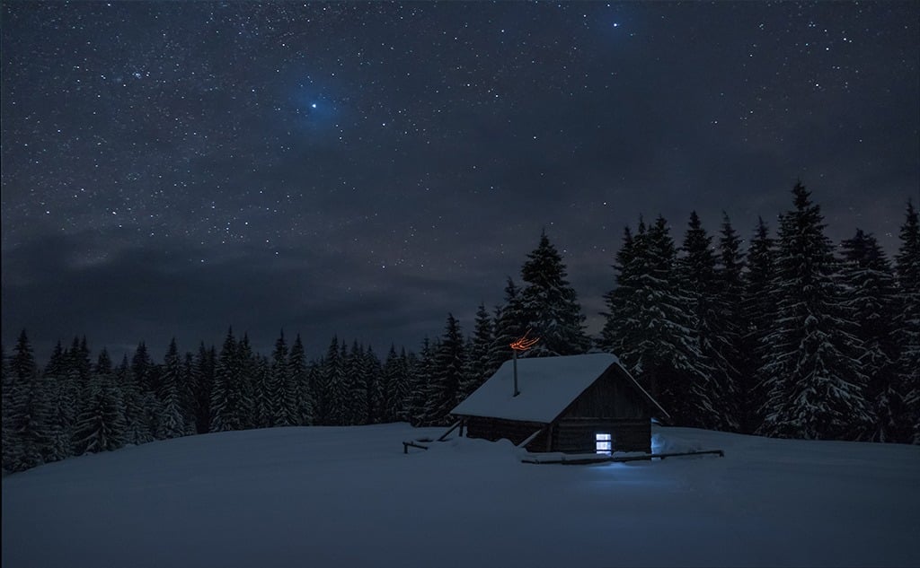 Wooden cabin under stars. Lights shines through the window from