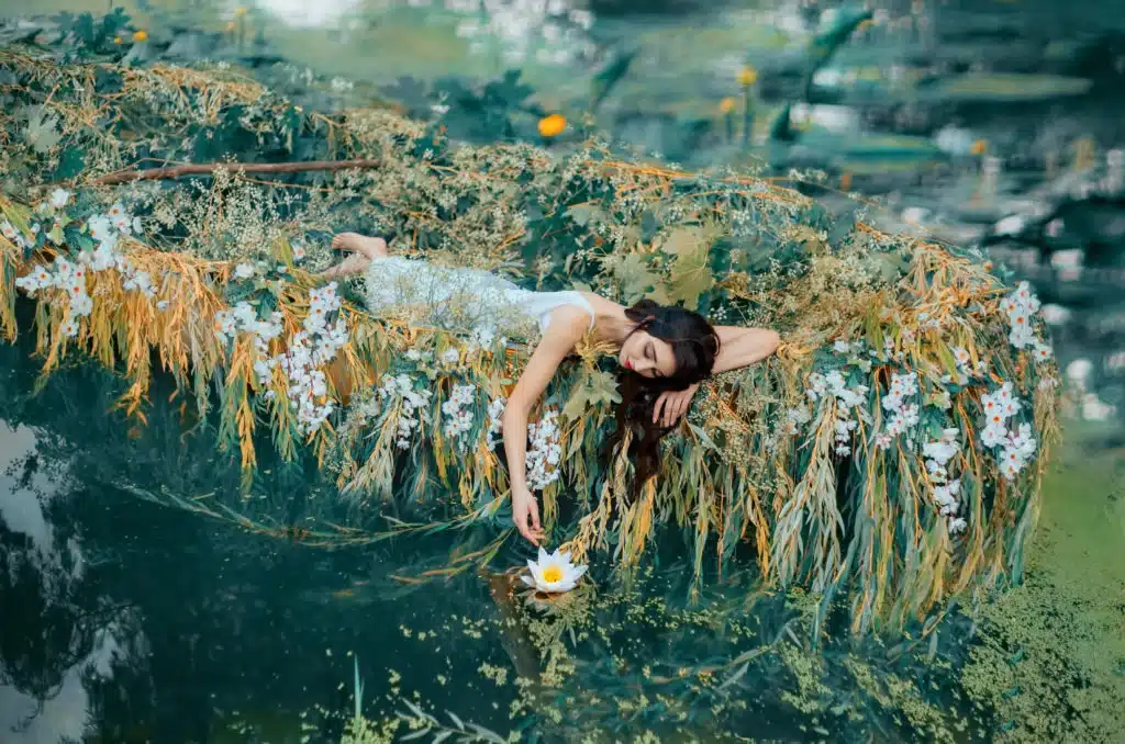 River Nymph in white vintage dress lies in boat decorated flowers yellow willow branches, enjoying silence and relaxing.