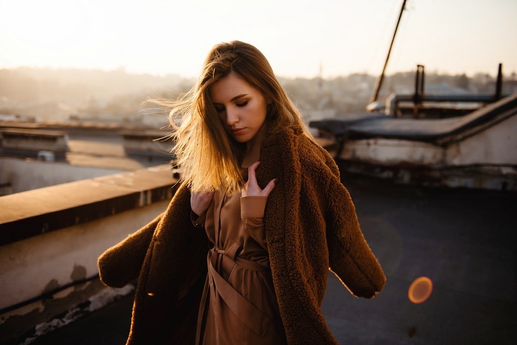 Attractive young brunette woman in stylish coat on the rooftop with beautiful city view on sunset.