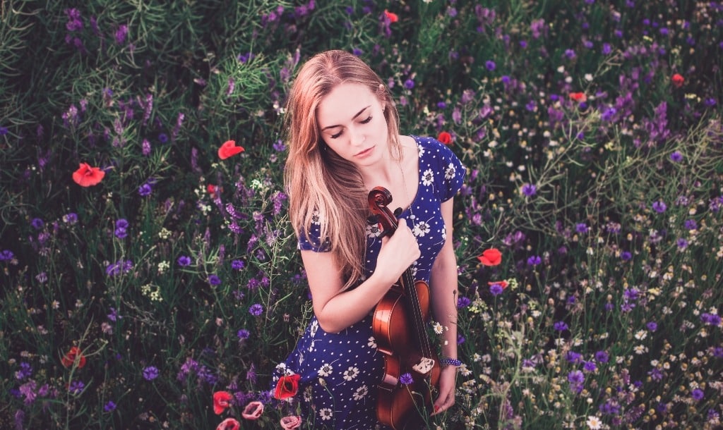 A young blonde girl holds a violin in the garden, with eyes closed.