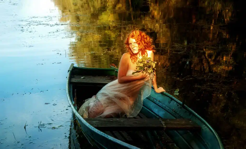 A young sexy red-haired woman sits in a boat like a siren in an old pre-Raphaelite painting.