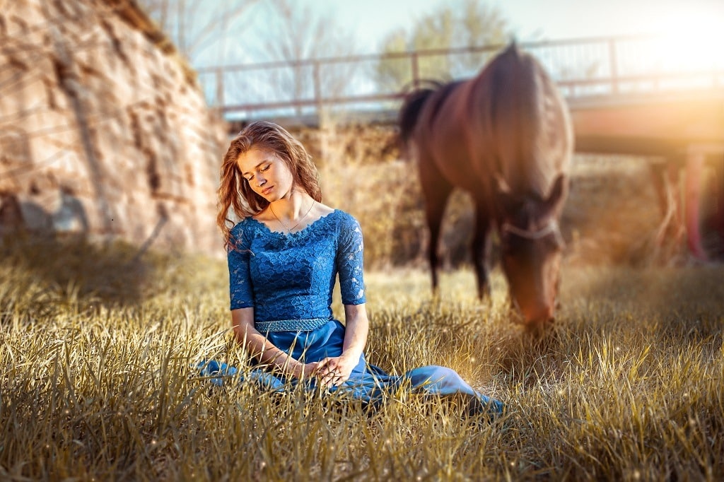 Attractive woman in blue dress seated on the grass, a brown horse behind her.
