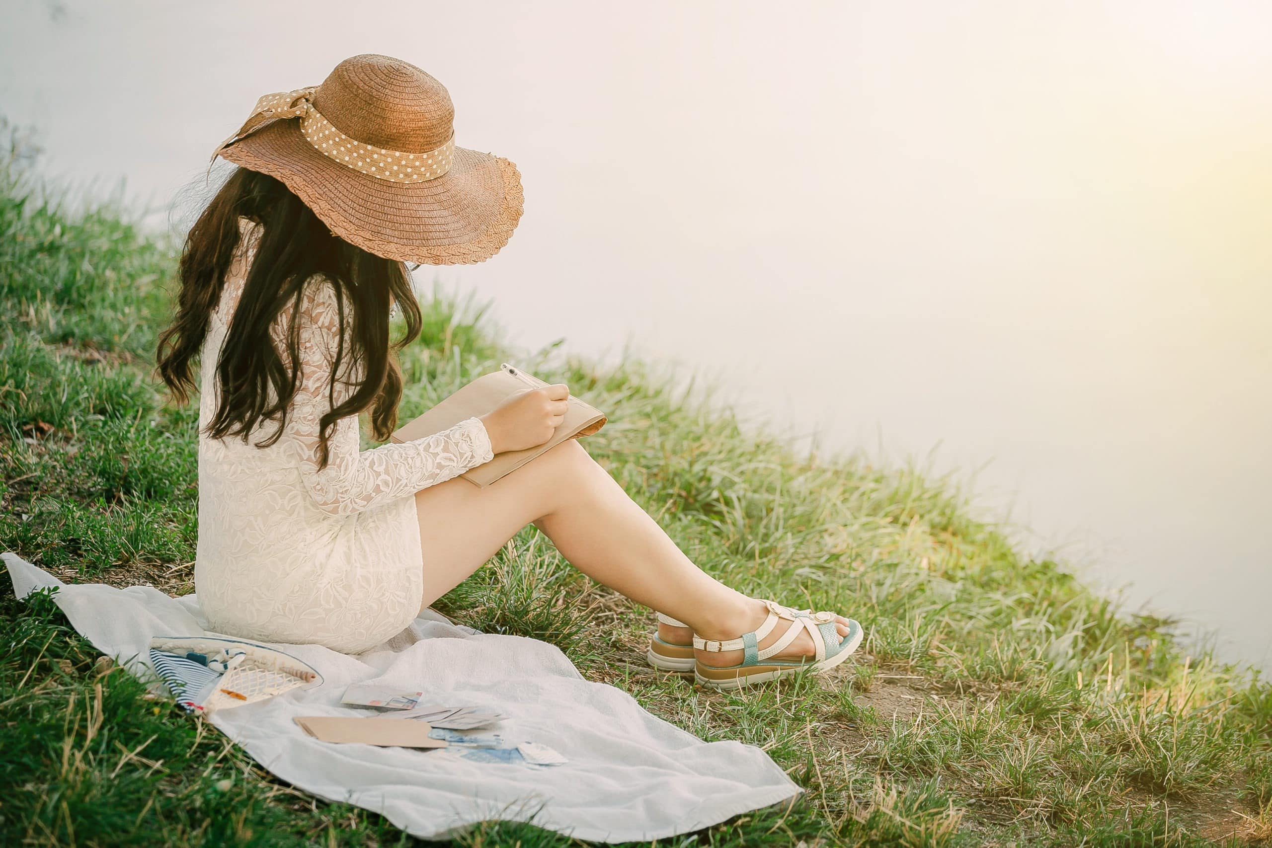young woman in a straw hat with long hair sits on the shore of the lake and writes