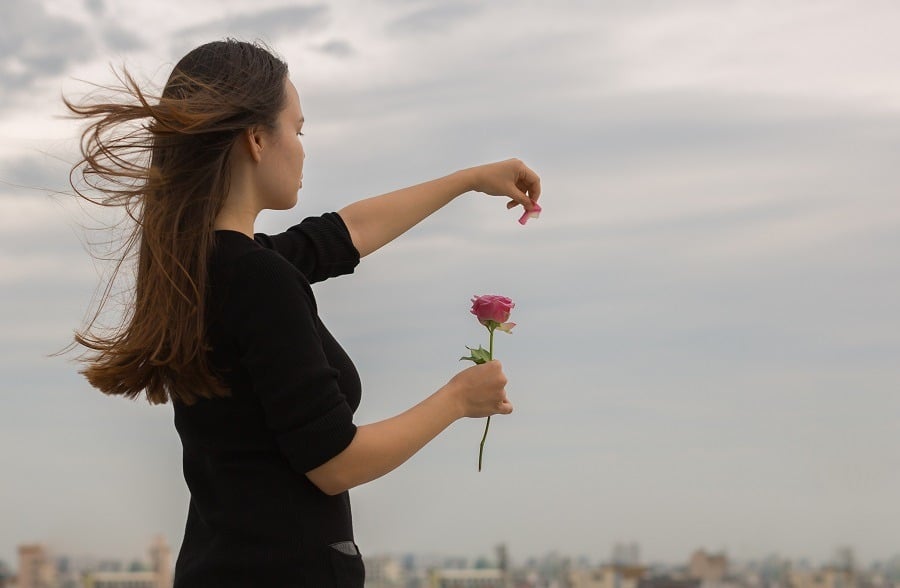 Young girl in black plucks petals from a rose lets them go into the air.