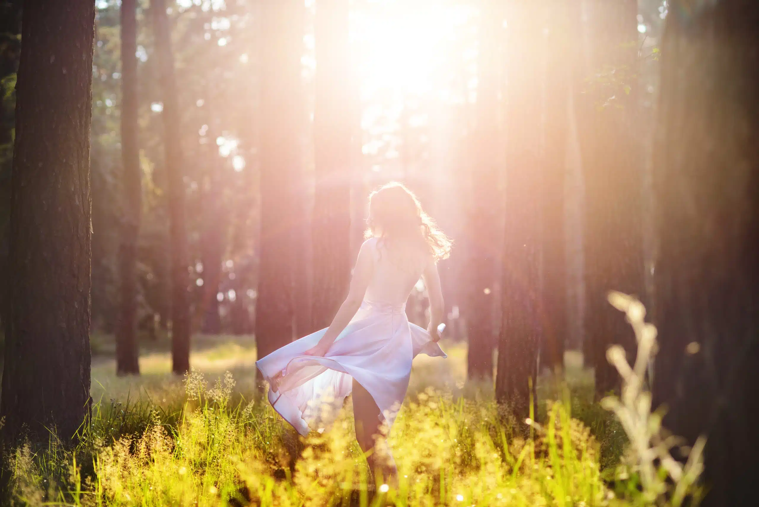Beautiful young woman wearing elegant light blue dress standing in the forest with rays of sunlight beaming through the leaves of the trees.