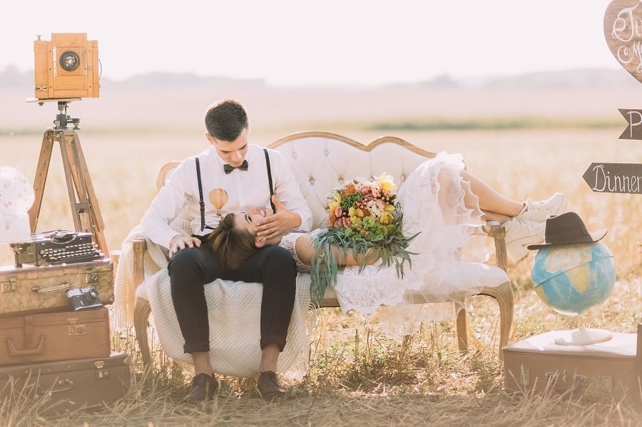 Smiling bride with colourful bouquet is lying on the lap of her vintage dressed groom at a summer field.