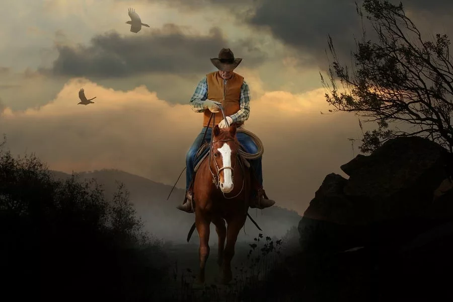 A mountain cowboy rides to the peak of a mountain with a beautiful cloudy sunset.