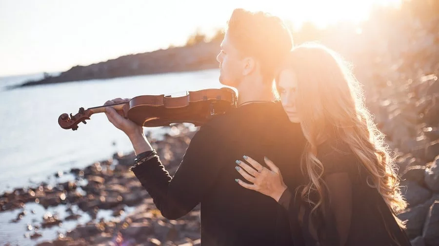 Girl hugging man from back as he plays the violin at seaside.
