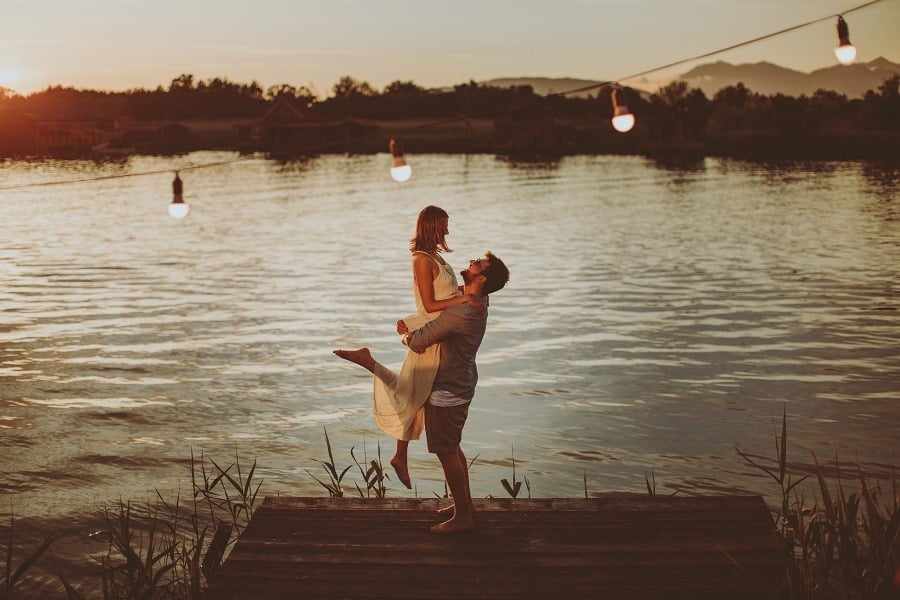 Man holds woman lovingly in the air by the river at sunset.