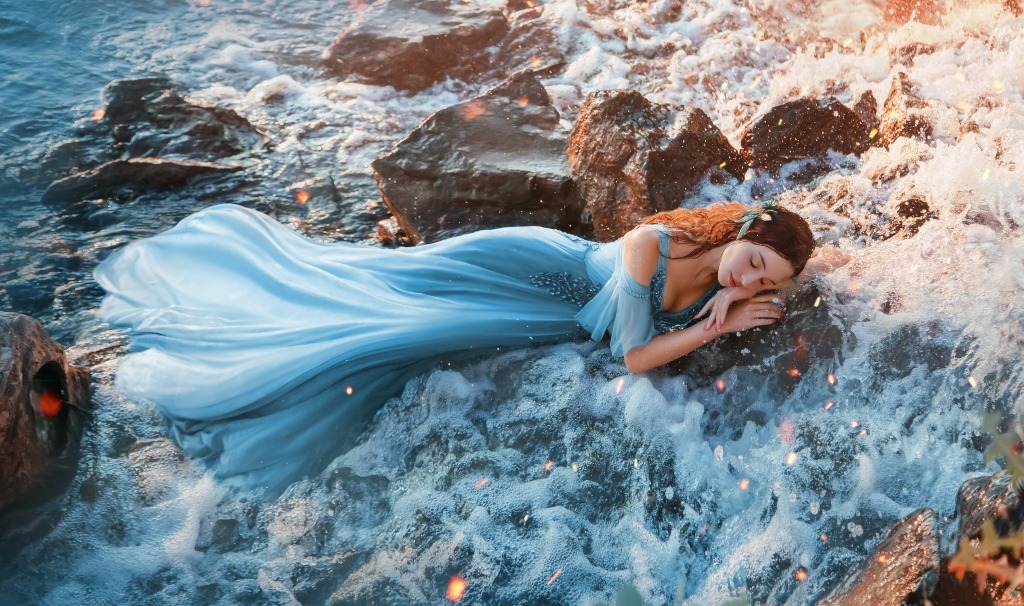 Charming sea princess resting on ocean stones, frothy water in the background.