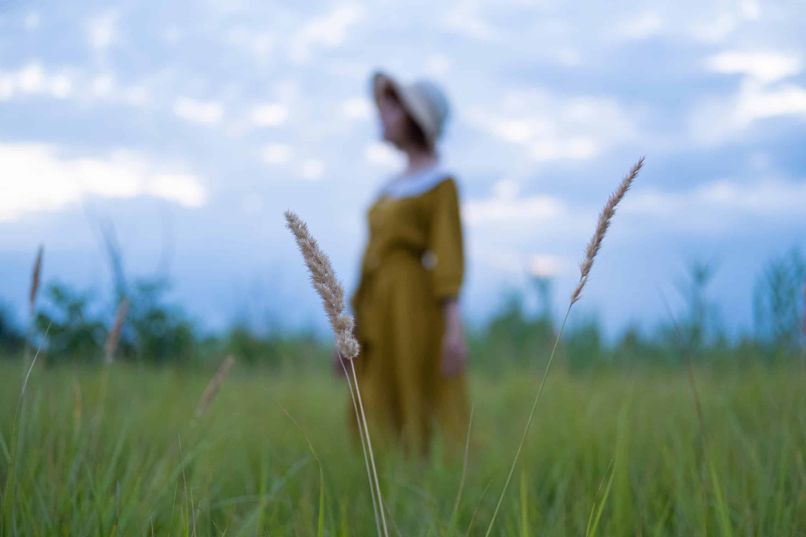 Melancholic woman wearing a dress and sunhat standing in the middle of grass field.