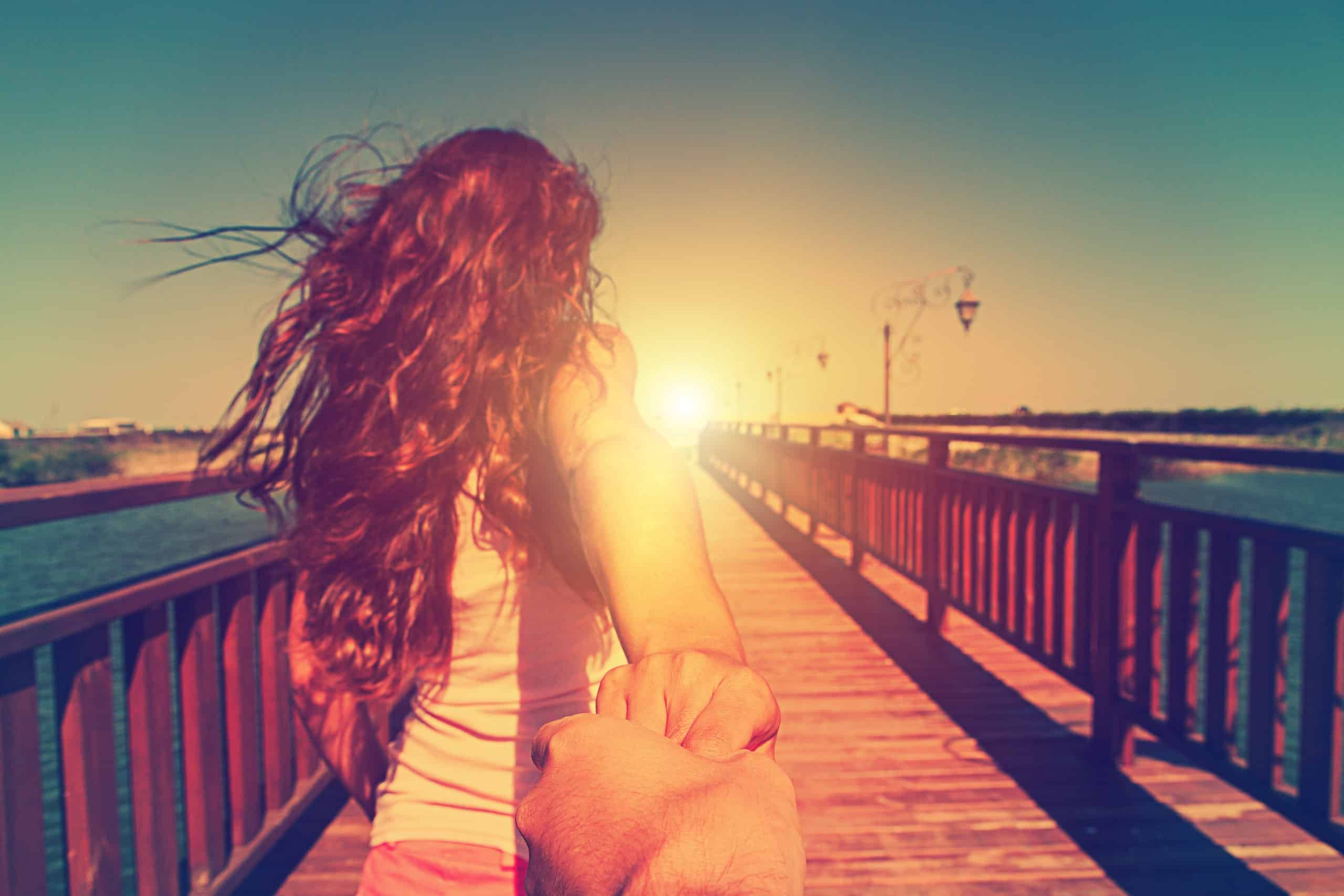Back view of young girl running toward the sunset on a bridge, holding a man's hand.