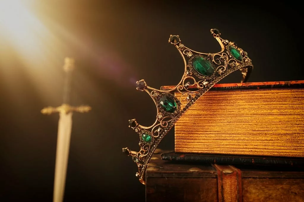 A king's crown over antique book next to a sword.