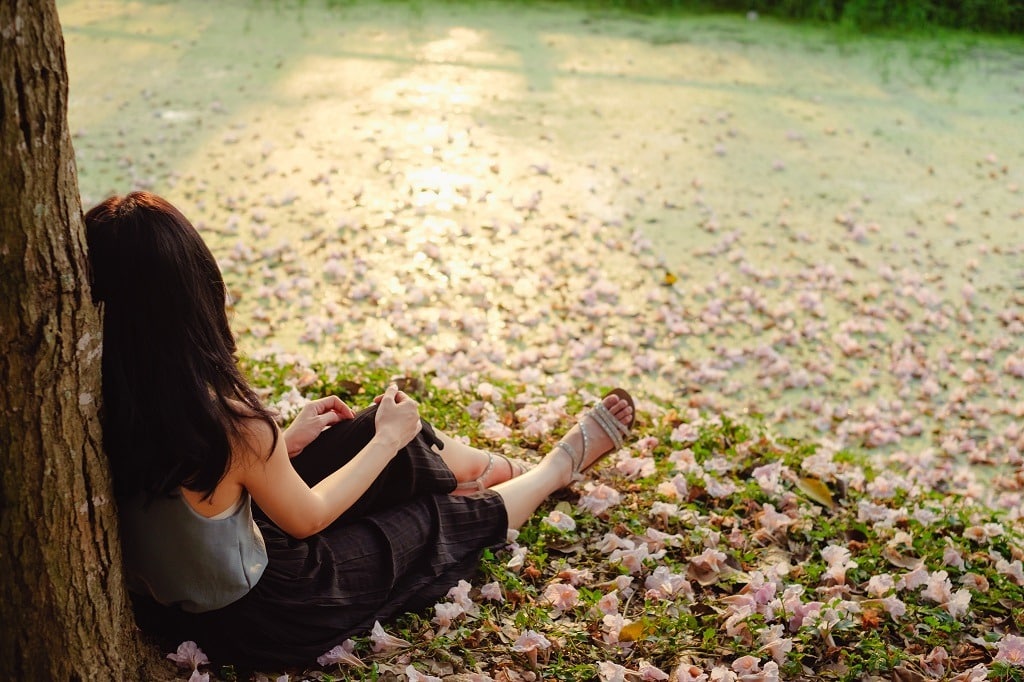 Sorrowful young woman sitting alone on green grass with falling pink flower.