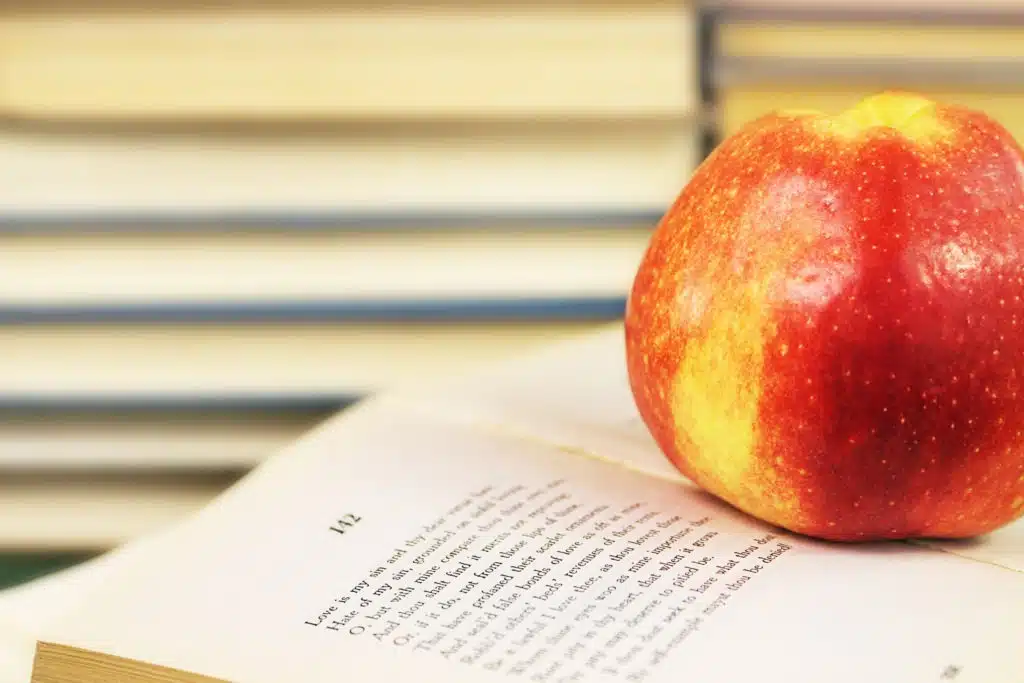  A large red apple on an open book with Shakespeare's sonnets on the background of a shelf with books.