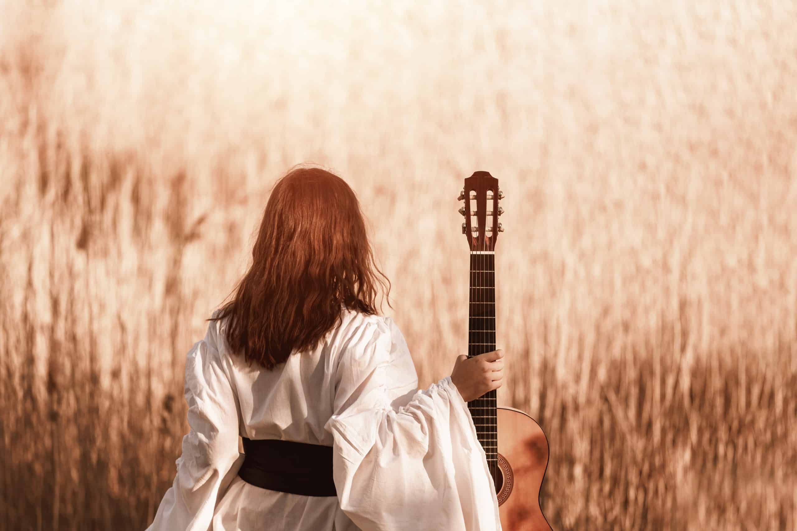 Beautiful young woman with red hair in a white medieval dress holding guitar and walking through the sunny field at warm light of sunset.