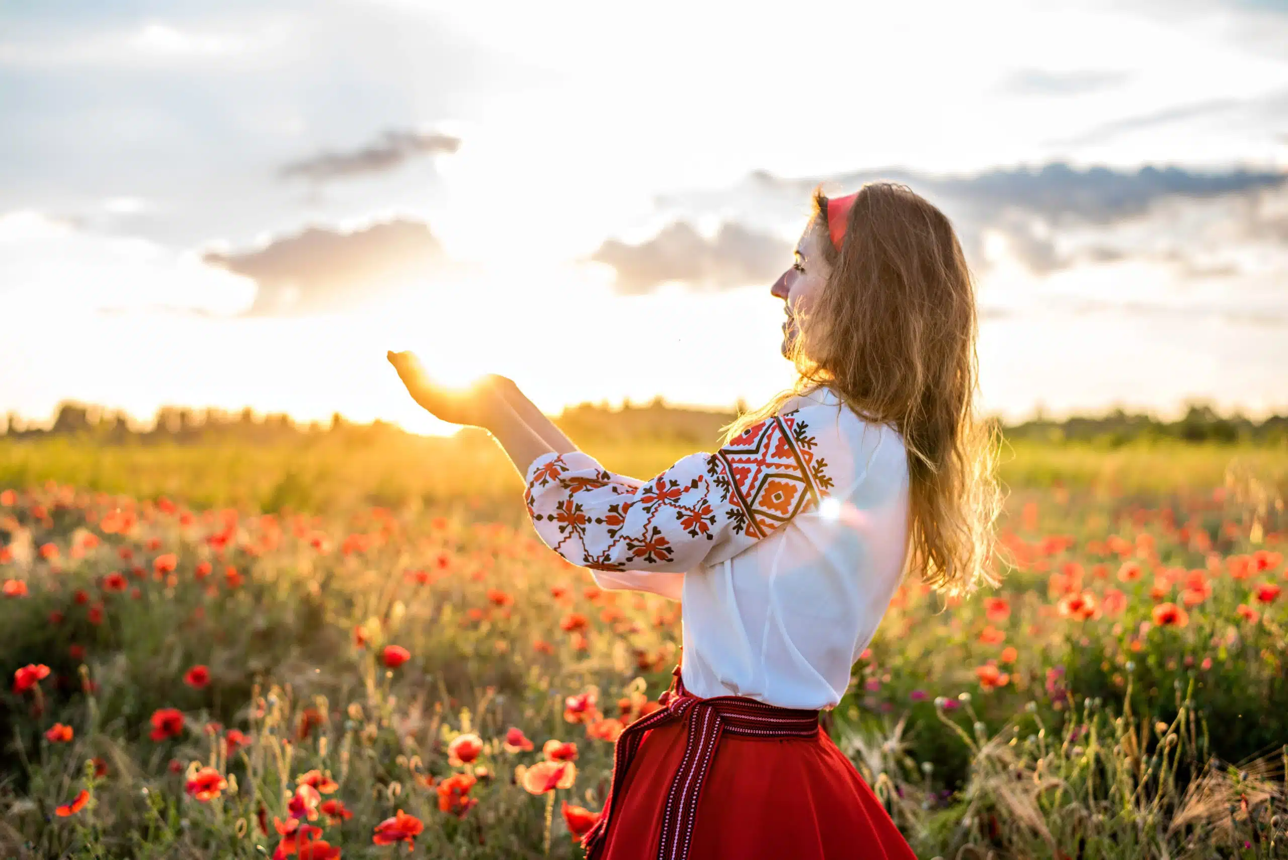 Woman in embroidery shirt holds the sun in hands in field of red poppies.