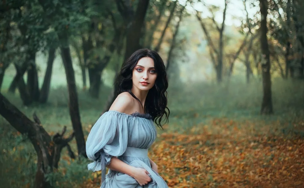 Lady with tar black hair and blue eyes, forest beauty in gray dress with bare shoulders hiding alone, portrait of enchantress in green grove with autumn leaves