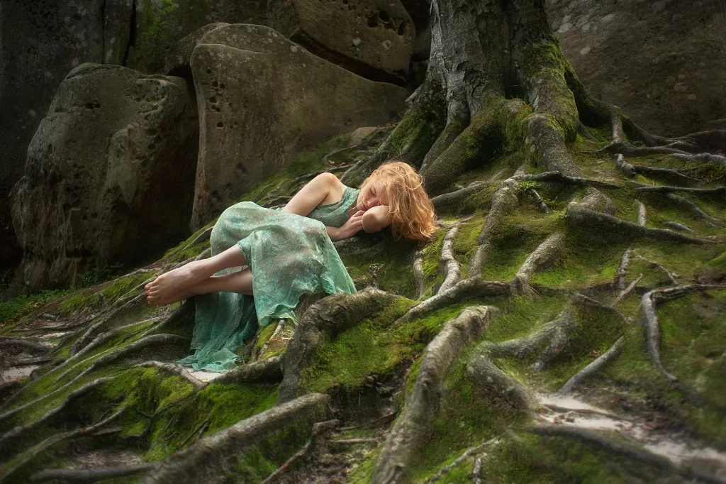 Sad red-haired woman in fairy forest on the moss and tree.