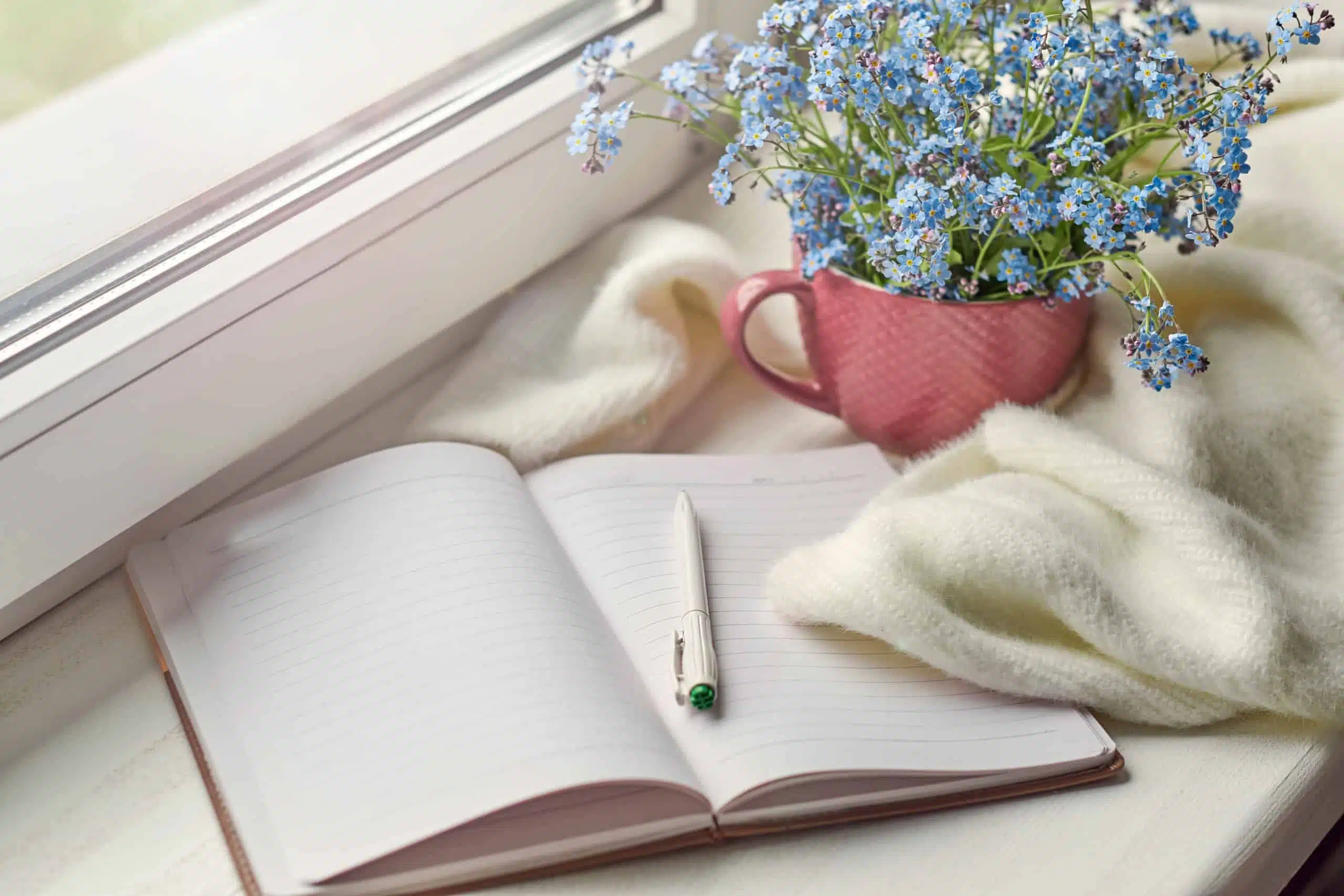 White warm plaid blanket, notebook and pen, flowers in cup by the window.