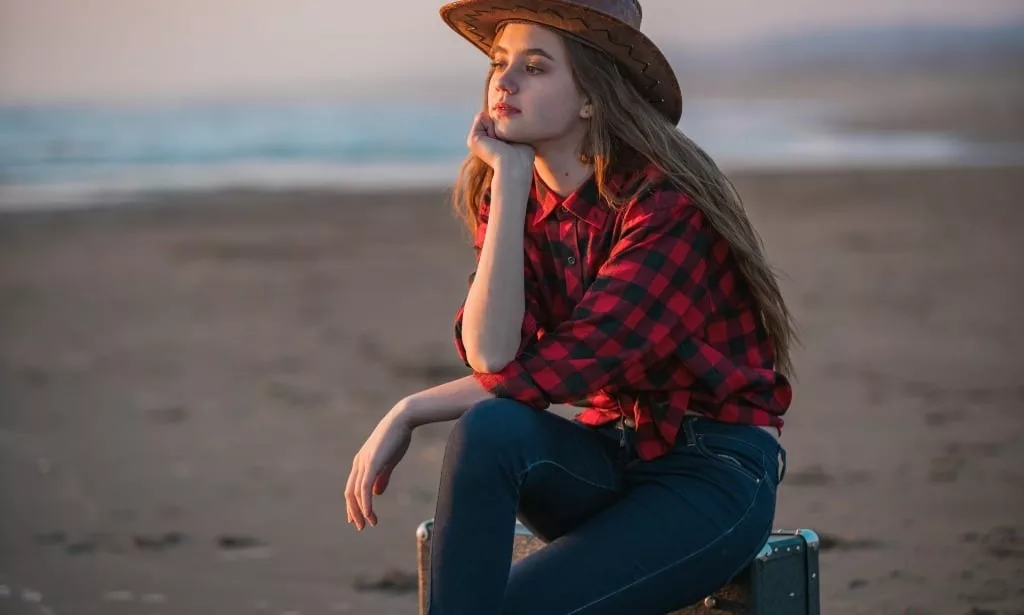 Young girl in a hat and red shirt sitting on luggage on the beach.
