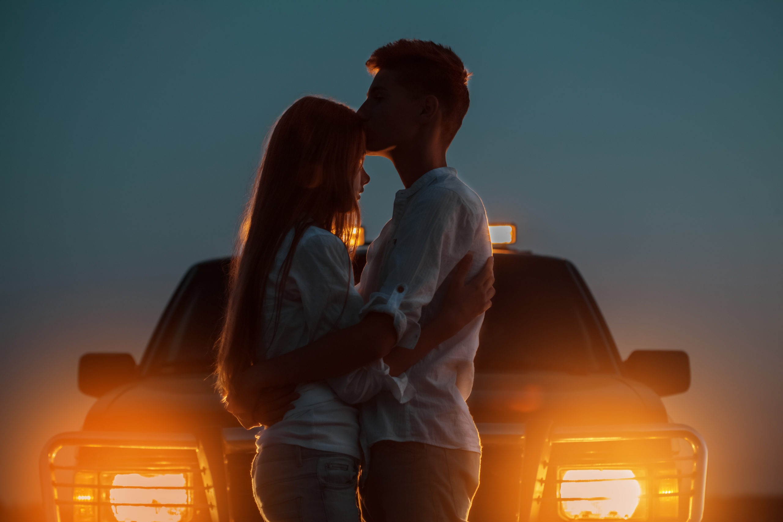 Silhouettes of young couple kissing goodbye in front of car headlights.