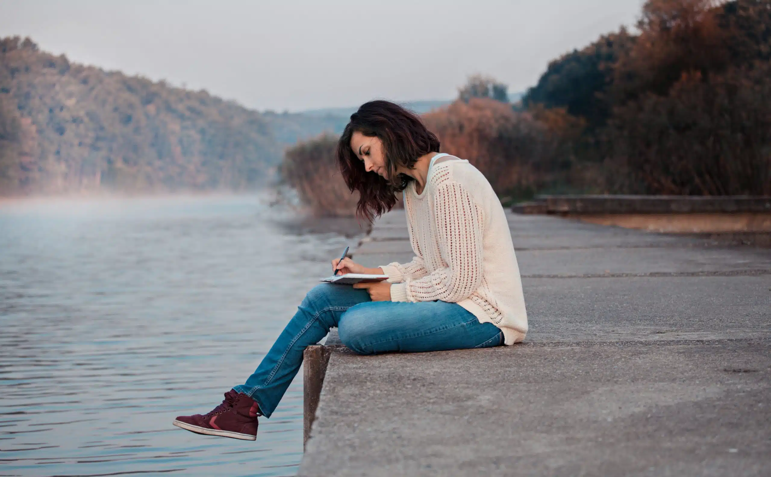 Woman in pensive mood seated at a dock writing on her notebook.