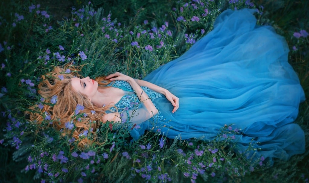 Stunning lady in blue gown lying in grass with purple blooms hugs herself with eyes closed.