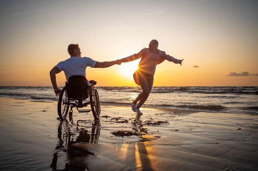 Handicapped man in wheelchair and his girlfriend holding hands on a beach at sunset.