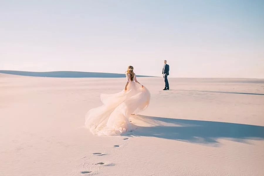 A woman in beautiful white gown runs to meet her man in the desert at sunset.