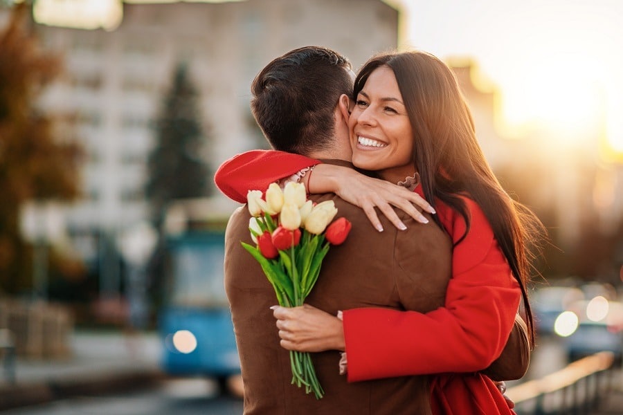 Happy woman holds beautiful bouquet in one hand, hugs partner on back view.