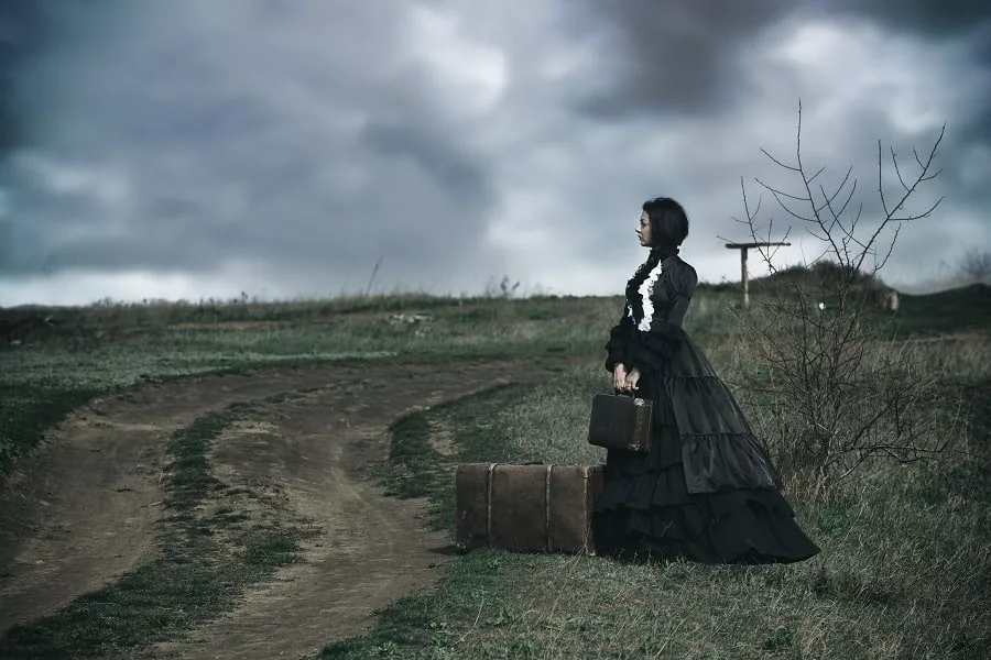 A victorian lady in black standing alone on the road with her luggage.