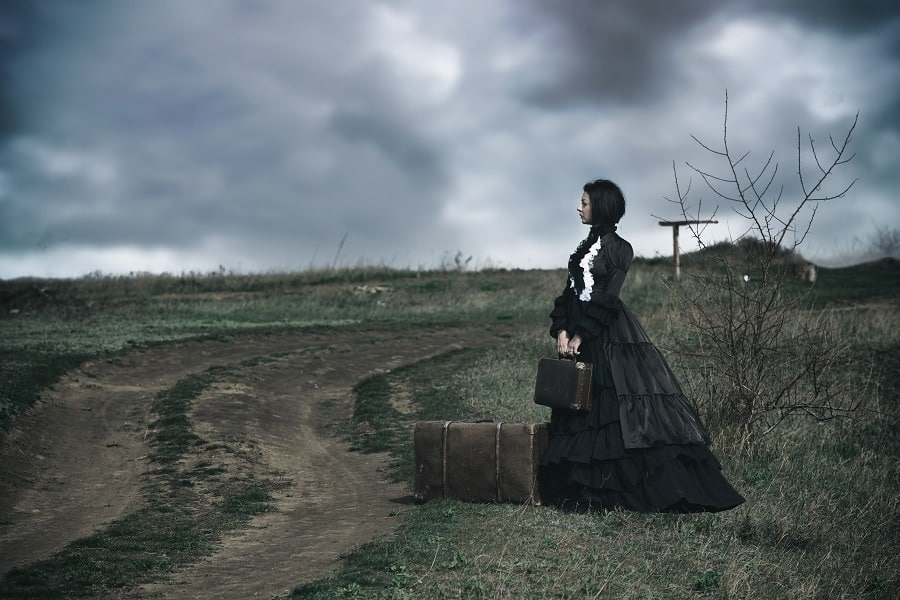 A victorian lady in black standing alone on the road with her luggage.