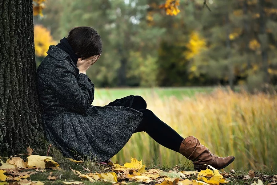 Woman hands covering face crying seated on the ground back to the tree in autumn.