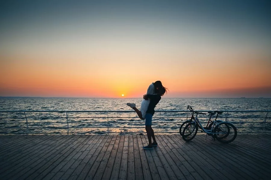 Man holding his girlfriend, kissing at the beach at sunrise sky at wooden deck in summer time.