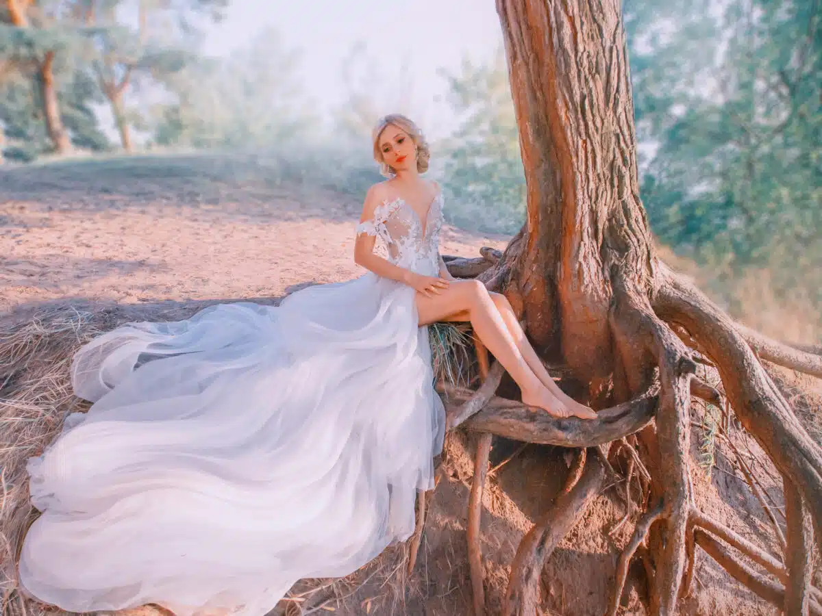 A mysterious, slender blond woman in a luxurious white dress. snow queen, sits near a tree with long roots. Fabolous forest. Hair collected elegant hairstyle. Stylish modern bride with attractive face